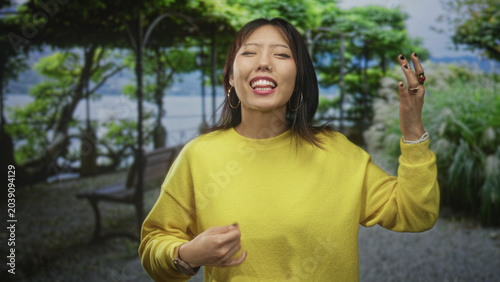 Valokuva Woman in yellow sweater checks watch and raises fingers by bench near lake in garden, grimacing with nose piercing visible; impatience