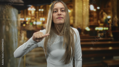 Foto Young hispanic woman pointing finger at camera and thumb to chest gesture in an ornate cathedral nave with wooden pews and warm lamps; confidence challenge