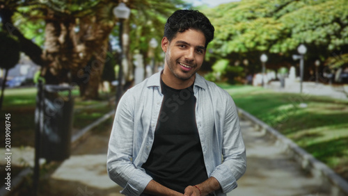 Konstfotografi Man smiling with hands clasped at waist on a tree lined street path, wearing light shirt over black tee; warmth contentment