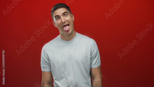 Schilderij op canvas Young hispanic man sticking tongue out showing tongue with tilted head in studio against red backdrop; playful mischief youthful energy