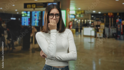 Schilderij op canvas Young woman wearing glasses biting her nails with hand to mouth gesture in an airport terminal near departure screens and queues; travel anxiety delay