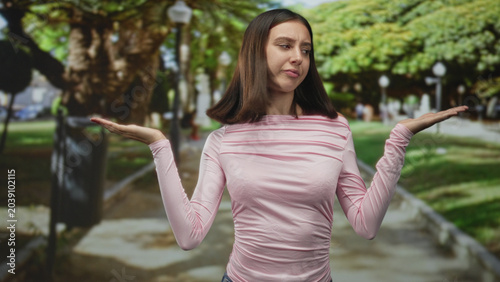 Valokuva Woman shrugging with palms up on a street park path, frowning expression and raised eyebrow; uncertainty choice dilemma