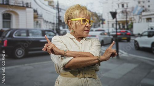 Cuadro en lienzo Woman senior hispanic with crossed arms and index fingers pointing outward, wearing yellow glasses and beige dress on a street near traffic and buildings; confidence assertiveness