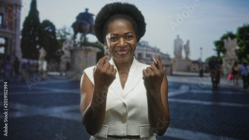 Photos Black woman smiling and showing middle fingers in a cobbled street plaza with statue and tourists, wearing white sleeveless vest; defiance confidence