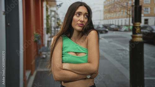 Schilderij op canvas Woman with bare shoulder in green crop top, arms crossed and pursed lips on street, urban sidewalk and parked cars visible; skepticism