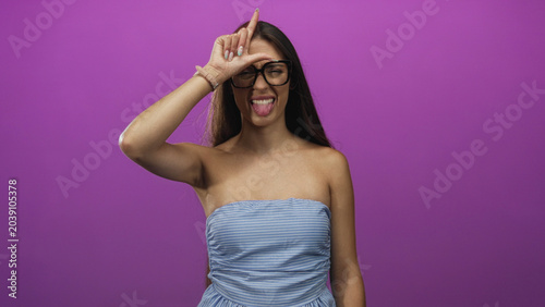 Slika na platnu Woman sticking tongue out and making l sign on forehead in purple studio wearing strapless blue dress; playful young fun