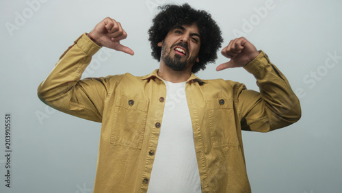 Bild auf Leinwand Man with curly hair wearing yellow jacket pointing thumbs to chest in studio with pale blue backdrop; confidence self pride