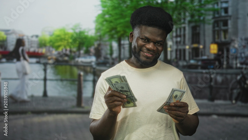 Canvas-taulu Man counting cash with hands while smiling on street by canal in amsterdam outdoors, holding us dollars in both hands; wealth success confidence