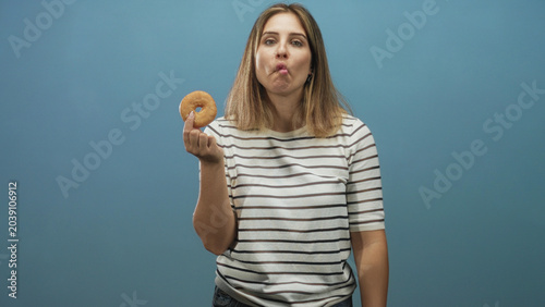 Картината върху платно Woman holding donut with fingertip and sticking tongue out while looking at camera against blue studio wall in a striped shirt; playful curiosity