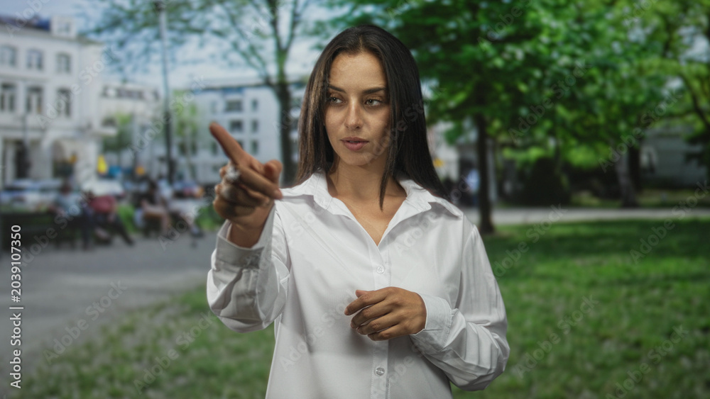 Fototapeta premium Woman points finger forward on city street near building, white shirt visible and fingertip aimed at camera; determination.