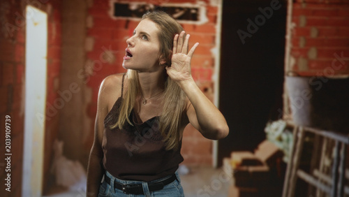 Photos Blonde woman cups hand to ear listening in an unfinished building amid red brick walls and a ladder, wearing a brown sleeveless top and jeans with belt; quiet curiosity