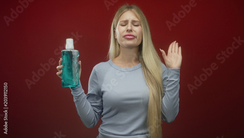 Fotografija Woman young blonde holding mouthwash bottle with eyes closed and grimacing, right hand raised in studio; disgust aversion oralcare