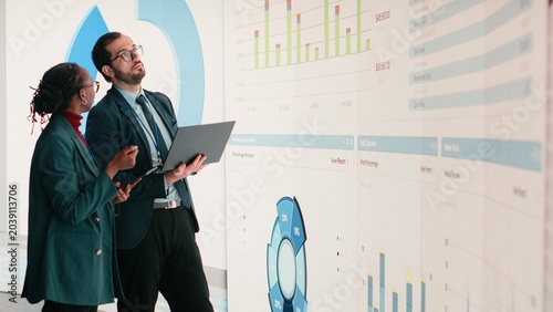 Obraz na plátně Corporate managers evaluate fiscal performance metrics on digital wall, collaborating, Diverse executive team discusses quarterly earnings report and shareholder equity charts during boardroom
