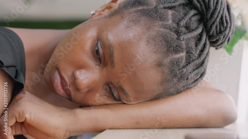 Black woman resting head on table, soft daylight, braided hair, tearful gaze. Postdate diner guest turned reflective home companion, pensive closeup capturing quiet vulnerability, cinematic grain,