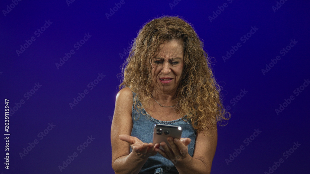 Obraz premium Middle age hispanic woman grimaces, holding a smartphone with both hands and squints at the screen in a studio; frustration technology challenge.