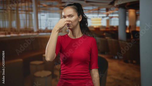 Billede på lærred Woman in red shirt pinches her nose and shields her face with one hand while gesturing in an airport seating area with lounge chairs and columns; disgust