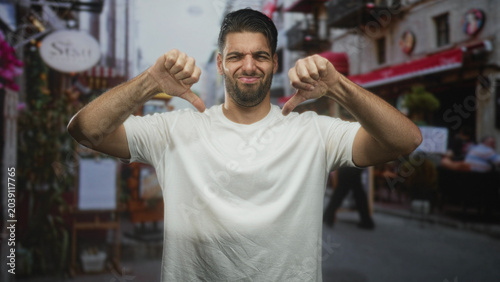 Vászonkép Man with beard shows thumbs down, bare thumbs visible, wearing white tshirt, standing and looking left in a street cafe with awning, menu board and outdoor chairs; disapproval critique