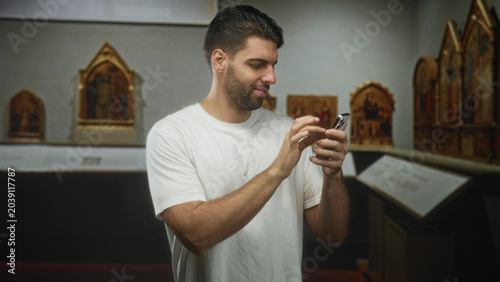 Fotografía Hispanic man with beard holding smartphone, tapping screen and pumping fist in building museum gallery with framed altarpieces visible; casual celebration joy