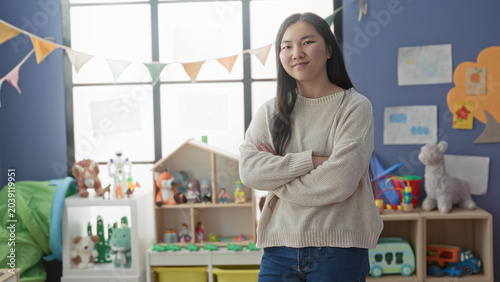 Εκτύπωση καμβά Young chinese woman with arms crossed beside a toy shelf and stuffed animals in a preschool building; calm nurturing