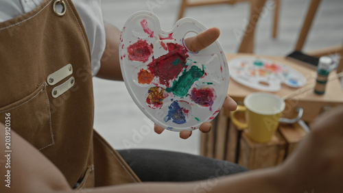 Photos Man painter holds paint palette and brush with fingertip through palette hole, mixing paint while seated at an easel in studio; concentration