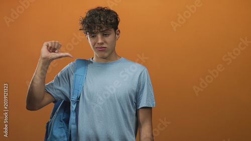 Bilde på lerret Boy wearing a backpack points finger left in a studio orange backdrop, grimacing then giving a thumbs down; disapproval rejection