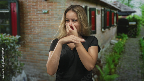 Schilderij op canvas Woman pinches nose at house entrance in front of a brick building, hand covering nostrils and gesturing with other arm outdoors; disgust discomfort