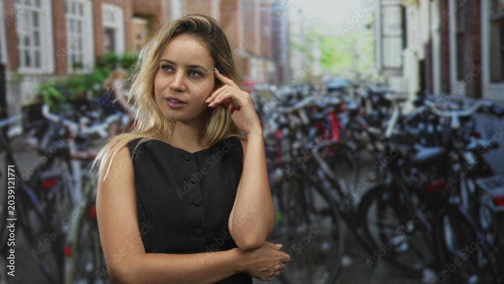 Fototapeta premium Woman wearing black dress with hand to forehead and arm folded across body on street in amsterdam; contemplative reflection.