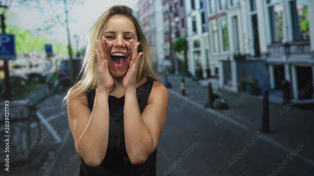 Fototapeta premium Young blonde woman cupping hand to mouth shouting on a narrow city street beside canal buildings and bollards, smiling broadly; excitement celebration.