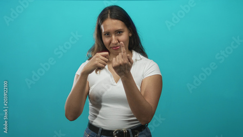 Vászonkép Young latina woman in white top shows middle finger to camera while standing in turquoise studio, casual jeans visible; bold defiance