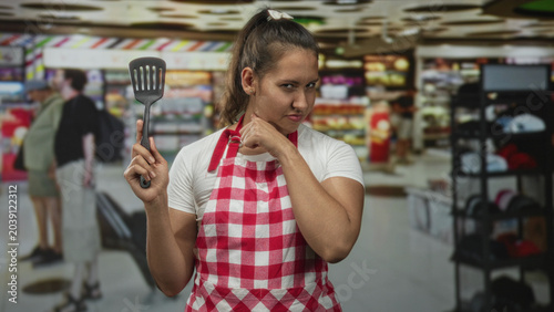 Konstfotografi Woman cook holding spatula points finger in busy airport terminal wearing red check apron and white tee, stern expression and raised hand; annoyance warning