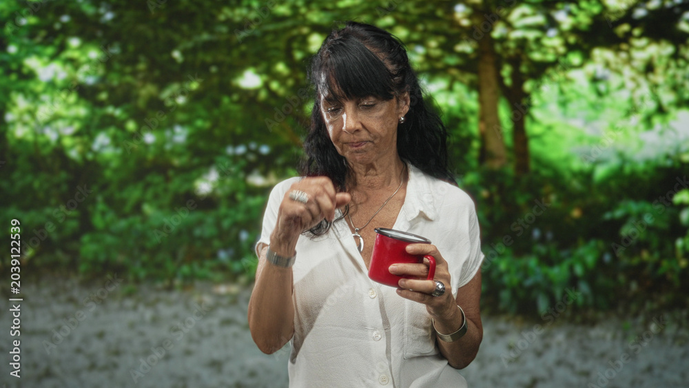 Obraz premium Senior hispanic woman in white blouse holds a red coffee mug with her right hand, puckers lips and glances aside while standing among leafy trees in a forest; contemplation.
