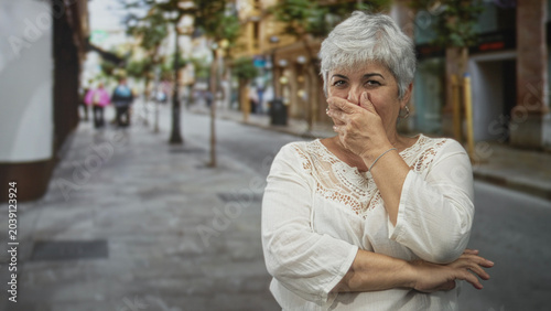 Tableau sur toile Woman plus size grey hair covering mouth with hand on street, arms crossed, eyes squinting in a suppressed laugh; amusement