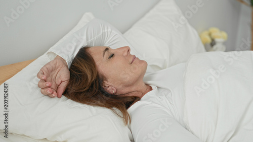 Photos Woman lying supine in bed with arm over forehead, hand near temple, eyes closed, pillow and white duvet, bedside plant and vase visible in bedroom building; calm rest