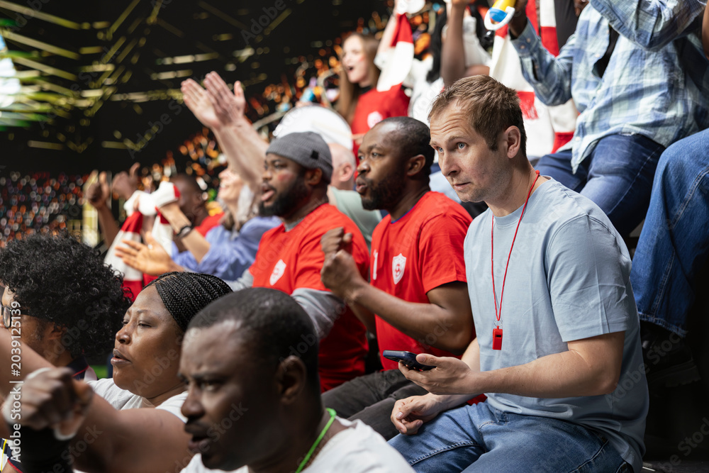 Fototapeta premium Soccer fan using betting app at the stadium during dramatic football match filled with cheering and joy. Supporter monitor live odds on gambling app, tension of sports betting and live match action.