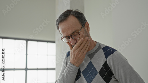 Canvastavla Man with hand on throat coughing in building interior, wearing glasses and argyle sweater; illness discomfort