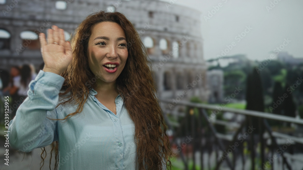 Fototapeta premium Smiling woman waving her hand at the coliseum building on an outdoor terrace railing near ancient arches and trees; travel joy.