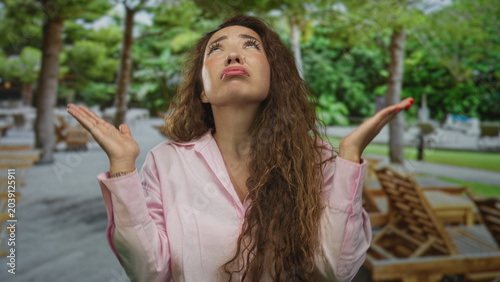 Foto Woman arms crossed pouting lips in park with wooden loungers and trees, hands with red nails; annoyance disapproval