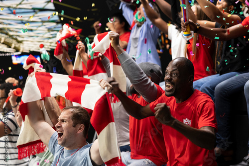 Tableau sur toile Excited football supporters celebrating with confetti in stadium stands, waving red scarves and cheering during soccer match victory and team spirit