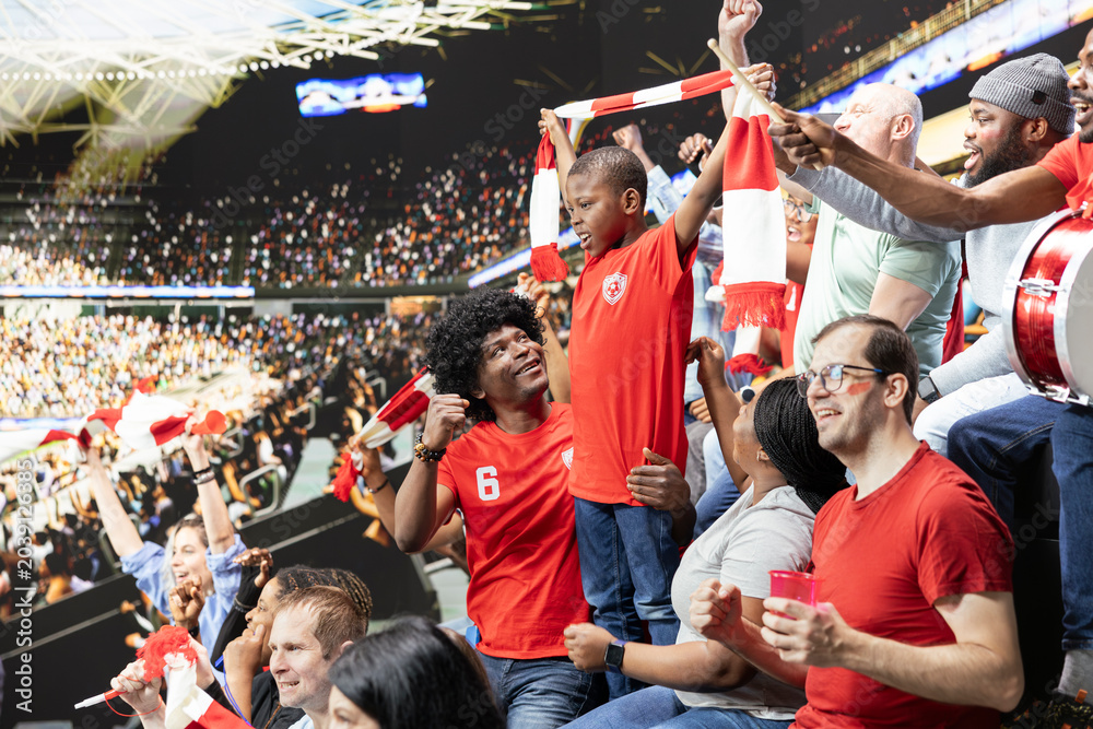 Fototapeta premium Excited parents and their little boy enjoy live football match together, surrounded by stadium crowd energy and passionate supporters. Black family sharing family outing with cheering.