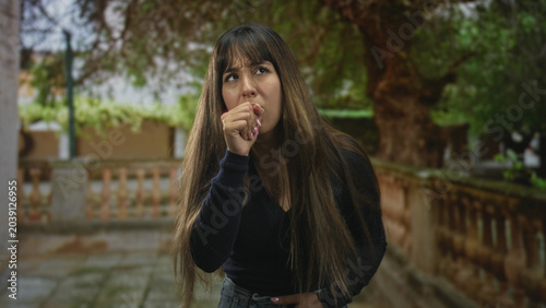 Foto Young woman with long hair in black sweater coughing into fist with hand on chest in stone building courtyard; illness concern health symptom