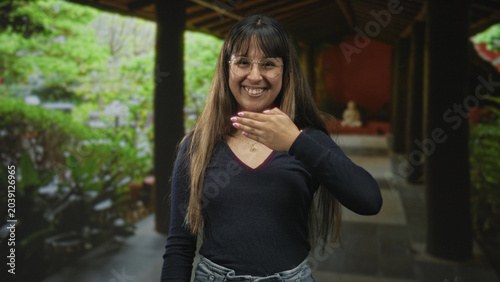 Photos Woman with hand to forehead and hand over chest, smiling and covering eyes in a covered wooden corridor building; playful greeting