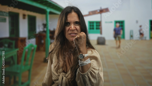 Foto Woman clenches fist on a street near a green building and cafe, wrist watch and rope visible; anger defiance resolve