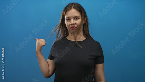 Foto Young caucasian woman in black t shirt points thumb over her shoulder while glancing sideways in a blue studio; annoyed