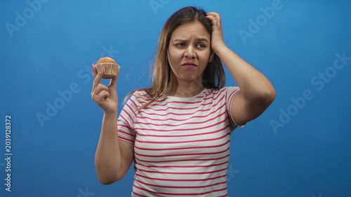Foto Young caucasian woman in pink striped shirt holding muffin with hand on head and grimacing in blue studio; disgust reluctance