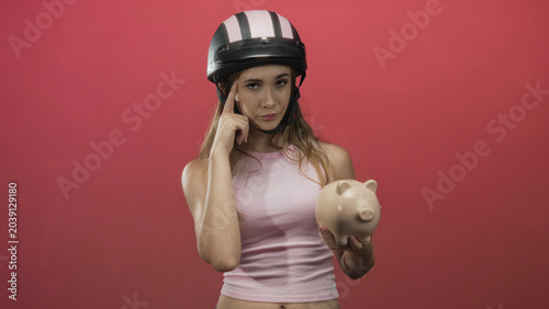 Photos Woman wearing helmet and sleeveless pink top holding piggy bank, finger pointing to temple in pink studio; financial concern