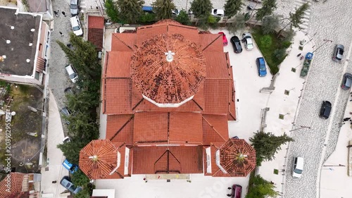 High-angle aerial tracking shot moving forward directly over the textured red-tiled roofs, central cross dome, and twin bell towers of the modern Orthodox Cathedral in Berat, Albania