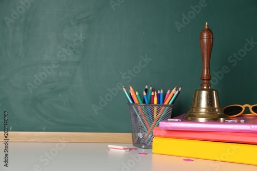 School holidays. Golden bell, sunglasses, notebooks and other stationery on white table near green chalkboard. Space for text