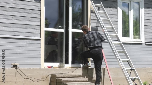 A man is renovating a wooden house, drilling a hole in the frame of a glass door outside the house