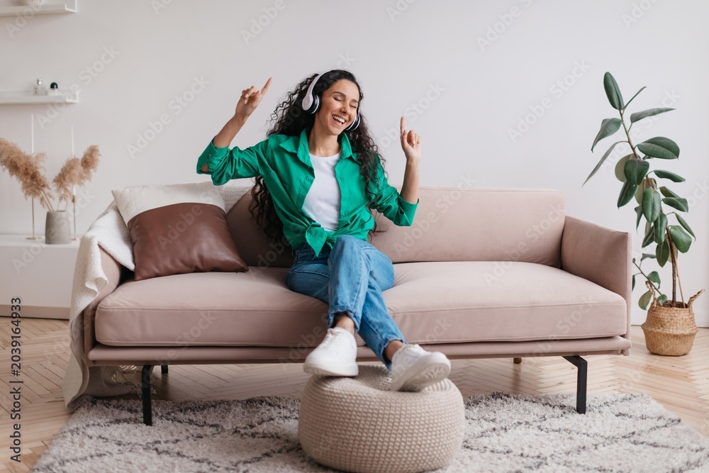 Obraz premium Smiling Young Woman Dancing And Enjoying Music While Sitting On Sofa In Cozy Living Room, Displaying Happiness And Joy In Indoor Comfort. Relaxed Home Lifestyle Concept.