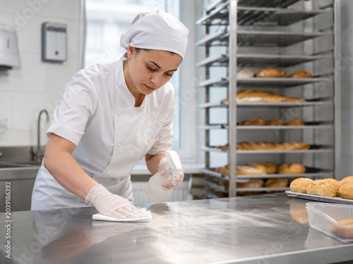 Bakery worker sanitizing a stainless steel worktable in a commercial kitchen with fresh bread racks in the background.